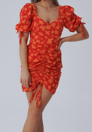 Kvinde iført en rød-orange blomstret kjole med puffede ærmer og en rynket front, der er bundet med justerbare snore ved kanten.