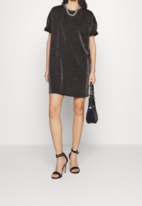 Robe noire à manches courtes avec un éclat métallique, dotée de manches retroussées et de fentes sur les côtés. Associée à des sandales à talons hauts noires avec des lanières.