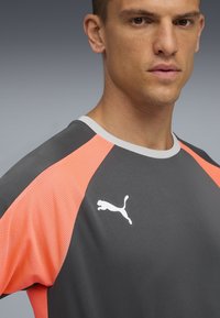Camiseta deportiva gris con mangas de malla naranja, logo de Puma en blanco, diseño de cuello redondo y un acabado texturizado.