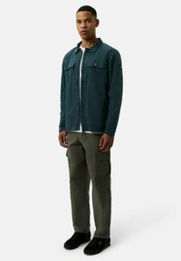 Zielona, lekka koszula zapinana na guziki z dwiema kieszeniami na piersi, w zestawieniu z oliwkowymi spodniami cargo z bocznymi kieszeniami i czarnymi trampkami.