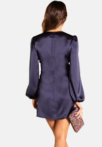 Robe mini en satin bleu marine avec des manches longues bouffantes, une couture arrière élégante et un ourlet arrondi. Le mannequin tient une pochette colorée et scintillante.