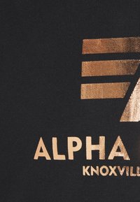 Tela negra con texto y diseño metálico en color dorado. "ALPHA" se muestra de forma destacada con una fuente block y un logo estilizado encima.