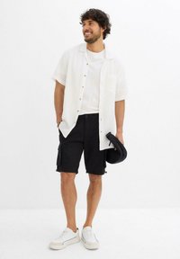 Camicetta bianca a maniche corte con bottoni, t-shirt bianca semplice, pantaloni cargo neri con tasche laterali e sneaker bianche. Completo semplice e informale.