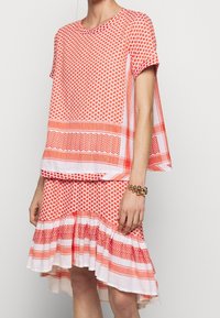 Žena v oranžovém a bílém vzorovaném topu s krátkým rukávem a odpovídající sukni s volánky, se zlatým korálkovým náramkem na pravém zápěstí.