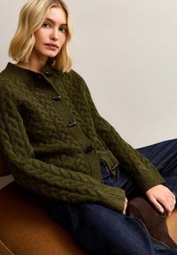 Dunkelgrüne Strickjacke mit Zopfmuster, hoher Kragen und schwarzen Knöpfen. Kombiniert mit Jeans und braunen Stiefeletten.