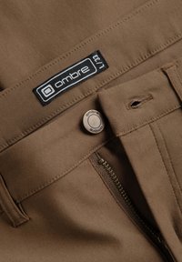 Pantalon marron avec fermeture éclair partiellement ouverte, bouton fermé et étiquette en tissu noire « ombre L/33 » sur la ceinture.