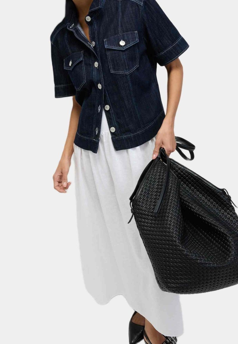 Personne portant une veste en jean sombre à manches courtes et une jupe blanche, tenant un grand sac à main noir tissé et portant des chaussures noires pointues.