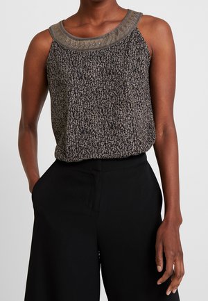 Vrouw die een mouwloos top met luipaardprint en kralen aan de hals draagt, gecombineerd met hoge zwarte wijde broekspijpen, staand met één hand in de zak.
