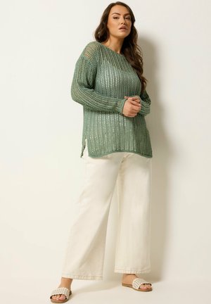 Frau trägt einen locker sitzenden grünen, locker gestrickten Pullover, weite weiße Hosen und beigen Slip-On-Sandalen, steht vor einem schlichten hellen Hintergrund.