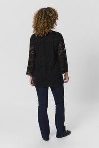 Black lace top with long, wide sleeves and scalloped hem, paired with dark blue flared jeans. Background is plain and light-colored.