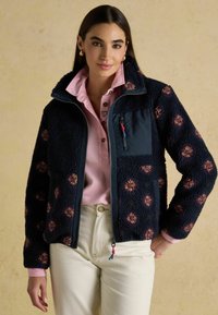 Marineblauwe fleecejas met bloemenpatronen, ritszakken en een hoge kraag. Draagt over een lichtroze shirt en gecombineerd met crèmekleurige broek.