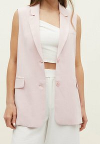 Ljusrosa ärmlös blazer med spetsig krage, två framknappar och sidofickor, kombinerad med en vit crop top och byxor.