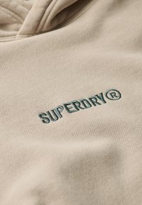 Sudadera beige hecha de una tela suave con un logo bordado de "SUPERDRY" en color azul verdoso. La textura es suave con un sutil patrón de grano.