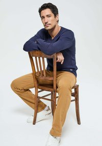 Un pull marin, un pantalon beige et des baskets blanches portés par un homme assis sur une chaise en bois ; le fond neutre met en avant les détails de la tenue.