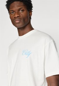 Camiseta de algodón blanca con un corte relajado, cuello redondo ribeteado y gráfico en cursiva color azul claro que dice "City" en el lado izquierdo del pecho.