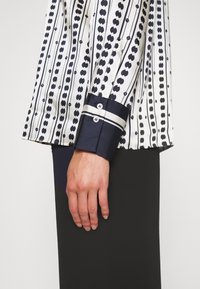 Chemise blanche avec un motif à pois navy, présentant un poignet navy foncé avec deux boutons et un accent rayé. Tissu lisse avec une coupe structurée.