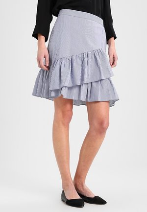 Femme portant une jupe à volants rayée bleu et blanc, des chaussures plates noires et un haut noir à manches longues, debout devant un fond uni.