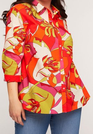 Kvinde iført en knap-til-op-skjorte med iøjnefaldende mønster med store blomster i rødt, orange, pink, grønt og hvidt, kombineret med blå jeans.