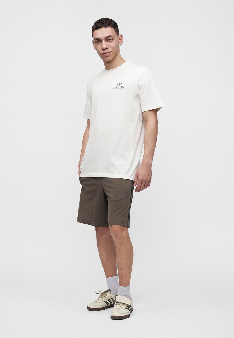Junger Mann steht und trägt ein weißes T-Shirt mit kurzen Ärmeln, olivgrüne Shorts, weiße Socken und weiße Sneakers mit schwarzen Streifen.