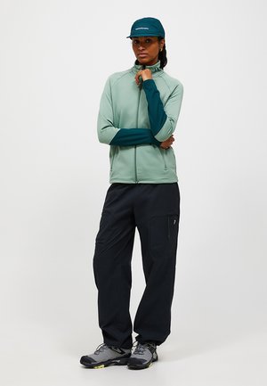 Kvinde iført en turkis lynlåstrøje, mørke cargobukser, vandresko og en turkis kasket, står med armene over kors i en neutral omgivelser.