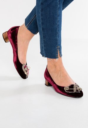 Voeten die donkerrode velvet blokhakschoenen dragen met versieringen van een juwelenhart en een gouden veiligheidsspeld, gecombineerd met versleten blauwe jeans.