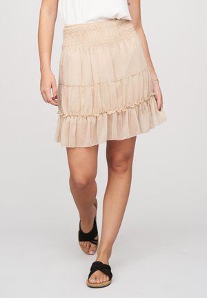 Vrouw draagt een beige gelaagde minirok met een gerimpelde tailleband, wit topje en zwarte sandalen met gekruiste bandjes.