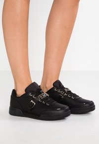 Baskets noires en matériau synthétique lisse, dotées de détails en doré, d'un emblème de marque et d'un col rembourré pour le confort.