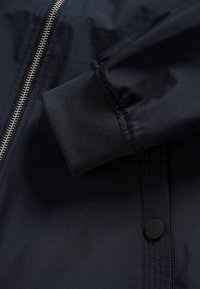 Marineblauw windjack met een rits, geribde manchetten en een drukknopsluiting. Gladde textuur met zichtbare stikdetails.