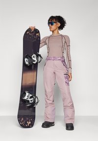 Snowboard mit einem dunkellila Untergrund und hellen Akzenten, kombiniert mit einem strukturierten, eng anliegenden Langarmshirt und hellrosa Schneehosen.