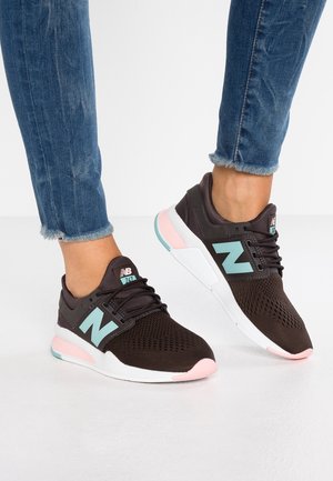 Fødder iført sorte New Balance-sneakers med turkise logoer og lyserøde detaljer, kombineret med blå jeans med frynset kant på et hvidt gulv.
