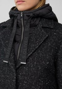 Vrouw in een gestructureerde donkergrijze jas over een zwart gewatteerde hoodie met rits en merkgebonden trekkoorden.