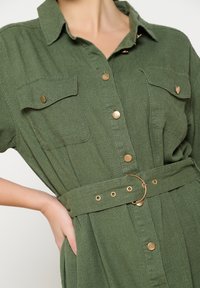 Roheline, lühikeste varrukatega shirt-dress tekstuuriga kangast, millel on kuldsed nööbid, kaks rinna taskut ja sobiv vöö ringikujulise kinnitusega.
