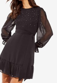 Svart klänning med genomskinliga långa ärmar prydda med spridda rhinestones. Har en figursydd midja och volang på nederkanten.