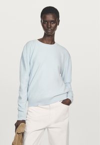 Ljusblått stickat tröja med rund hals, ribbit fåll och muddar, bärs med vita byxor. Texturerad tyg och avslappnad passform.