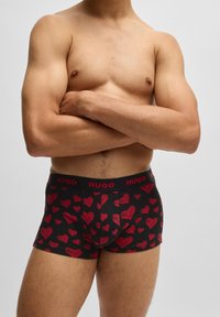 Bóxers negros con un patrón de corazones rojos, que presentan una cinturilla con el logo rojo "HUGO". El material parece suave, con un diseño ajustado.