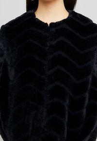 Veste en fausse fourrure noire avec un motif ondulé texturé et un col rond, offrant une apparence douce et pelucheuse, sans fermetures visibles.