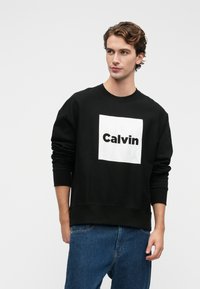 Čierna mikina so bielym obdĺžnikovým logom, na ktorom je výrazným písmom napísané "Calvin". Dizajn obsahuje rebrované manžety a spodný lem.