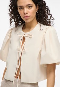 Kurzärmlige, cropped Bluse in hellem Beige mit Puffärmeln, Rundhalsausschnitt und einer vorderen Bindeschließe. Glatte Stofftextur.