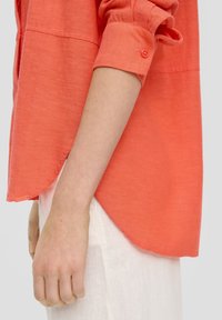 Koraal linnen shirt met opgerolde mouwen, voorzien van een knoopdetail en een gebogen zoom. Draag het over een witte, gestructureerde stof eronder.