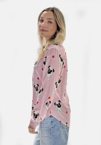 Langärmliges rosa Hemd mit wiederholtem Hundemuster und roten geometrischen Akzenten, kombiniert mit hellblauen Jeans. Weiche Stoffstruktur.