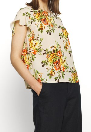 Mujer con blusa beige de manga corta con flores naranjas y amarillas, combinada con pantalones negros, manos en los bolsillos.