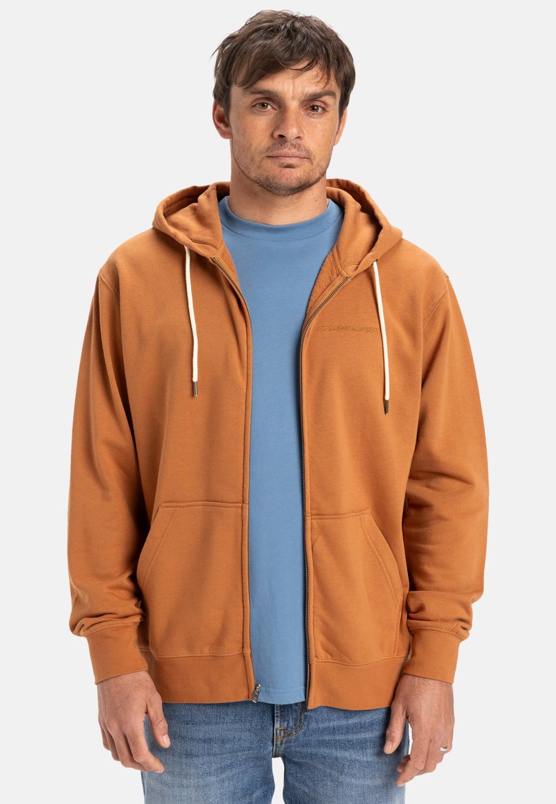 Rostfärgad zip-up huvtröja med en framficka, vita snoddar och ett lätt tyg. Bärs över en blå t-shirt.