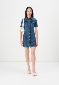 Robe en denim à manches courtes, avec un bouton sur le devant et un col. Couleur bleu foncé avec des détails de couture clairs, dotée de deux poches poitrine. Texture lisse.