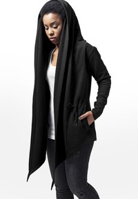 Cardigan à capuche noir en tissu doux, présentant un design asymétrique, des poches avant et un cordon de serrage à la taille.