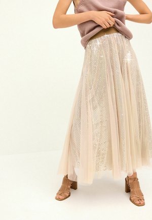 Femme portant une jupe longue crème transparente ornée de paillettes, des sandales à talons blocs nude à brides et un top en maille sans manches.
