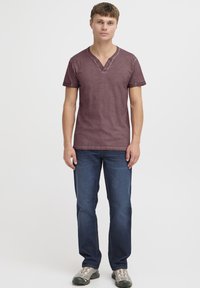 Burgunderfarget kortermet V-hals t-skjorte med tre-knaps placket, brukt sammen med blå jeans og grå sneakers, vist mot en nøytral bakgrunn.