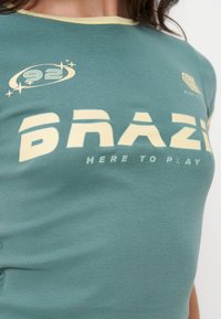 T-shirt vert avec texte jaune crème « 6NAZI HERE TO PLAY » et petits logos, porté par une personne visible du cou à la taille.