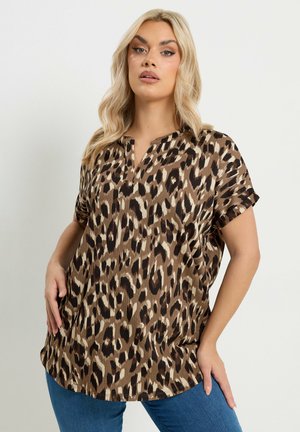 Blonde Frau, die eine kurzärmelige braune Bluse mit Leopardenmuster und V-Ausschnitt sowie eine blaue Jeans trägt, steht vor einem einfarbigen Hintergrund.