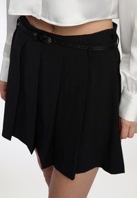 Gonna nera plissettata realizzata in tessuto liscio, con una sottile cintura nera con fibbia argentata, indossata con un blouse corto bianco.