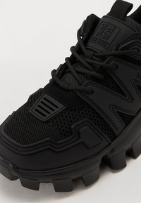 Chaussure de sport noire avec un dessus en mesh et en caoutchouc, accents texturés, languette rembourrée et semelle extérieure épaisse à motif. Logo sur le côté.